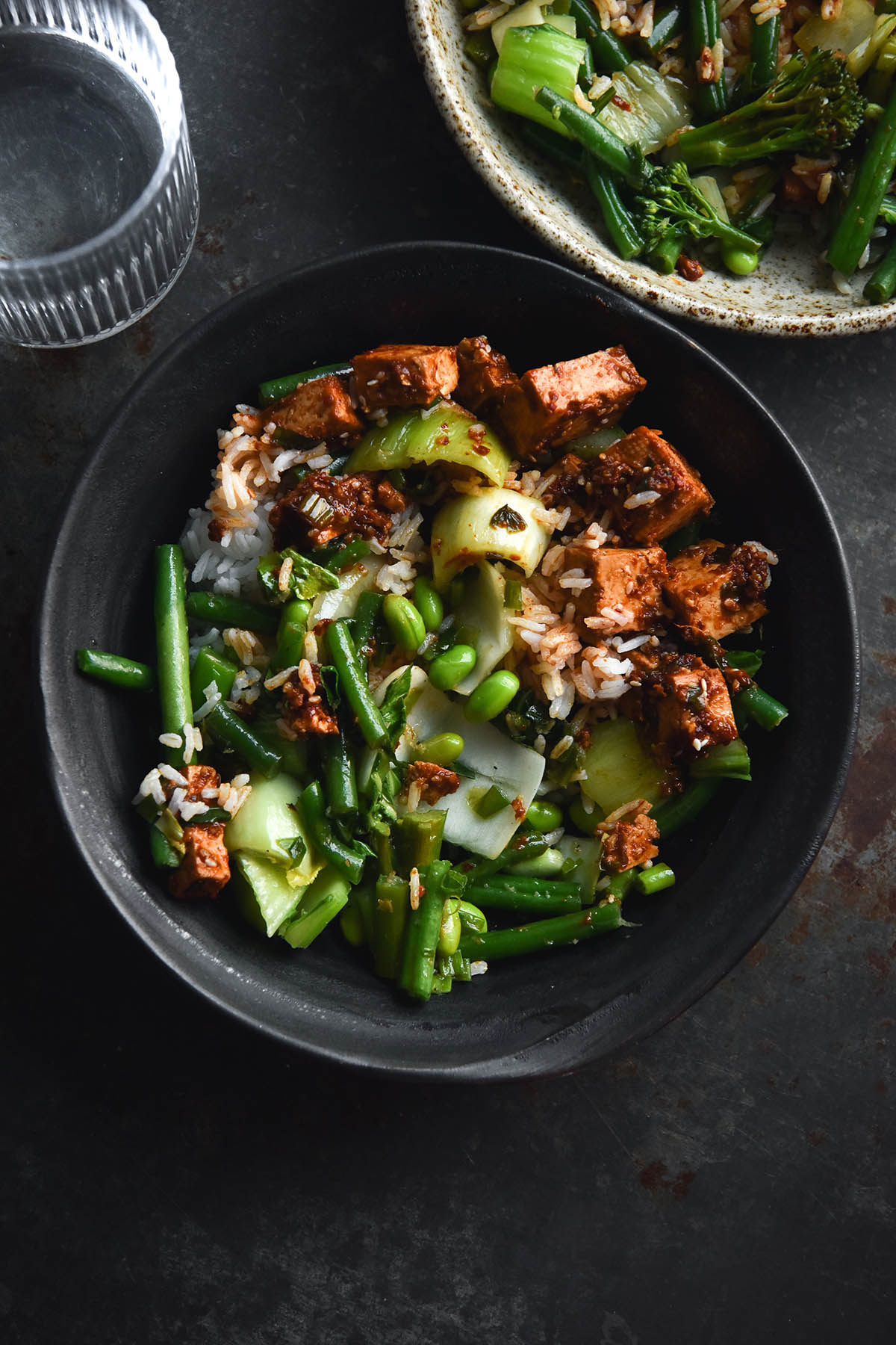 An aerial close up view of two bowls of chilli oil tofu, Asian greens and rice mixed together. The bowls are dark blue ceramic and the background is dark mottled grey. A glass of water sits to the top right of the image, one bowl sits in the centre of the image, and the other sits in top right hand side
